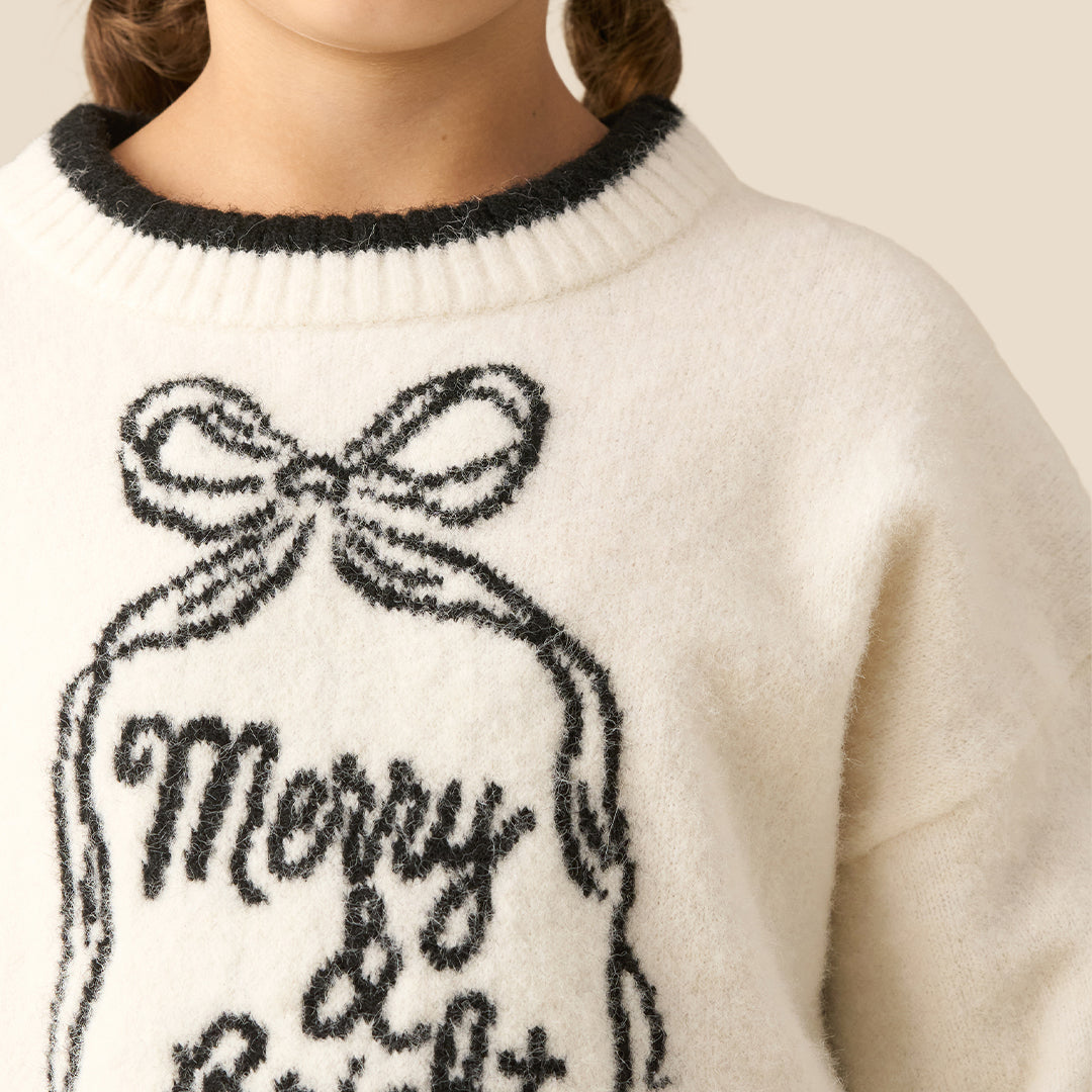 A young girl smiling while wearing a cream-colored knit sweater with a black 'Merry & Bright' ornament graphic and black trim, paired with black faux leather pants, against a solid beige background.