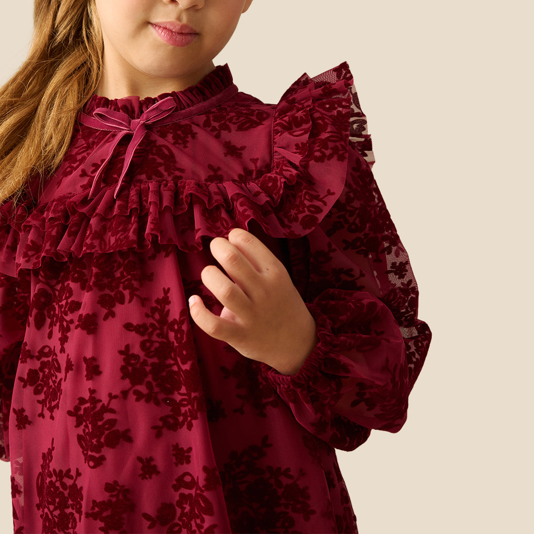 A young girl with her hair in a ponytail models a long-sleeved burgundy dress with a floral lace overlay, ruffled collar, and tiered skirt, set against a solid beige background.