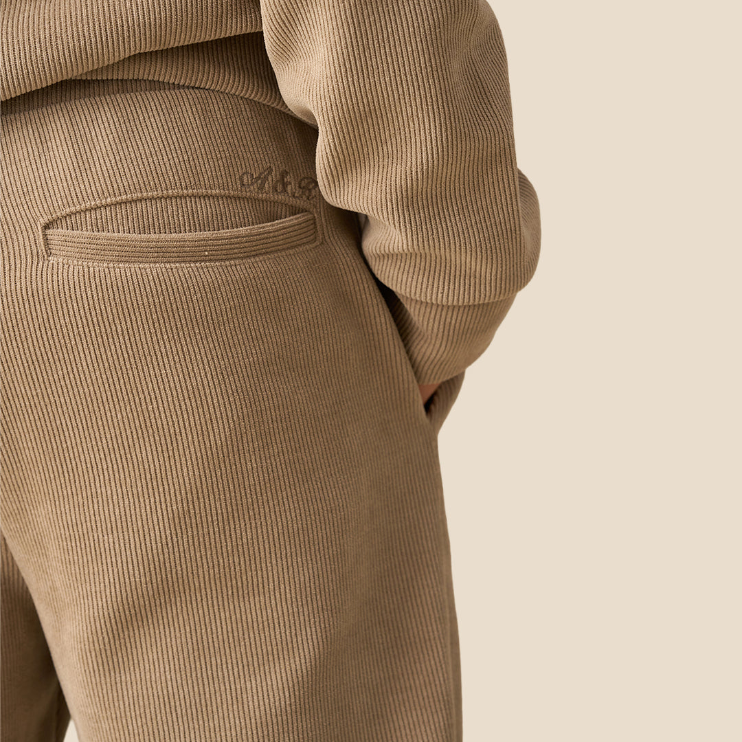 A child wearing a matching beige fleece tracksuit with a half-zip sweatshirt and relaxed-fit pants, paired with brown sneakers, against a solid light beige background.