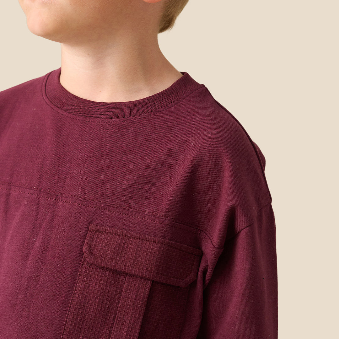 A young blonde boy wearing a long-sleeve crewneck shirt in a deep burgundy color, featuring a chest pocket, standing against a solid light beige background.