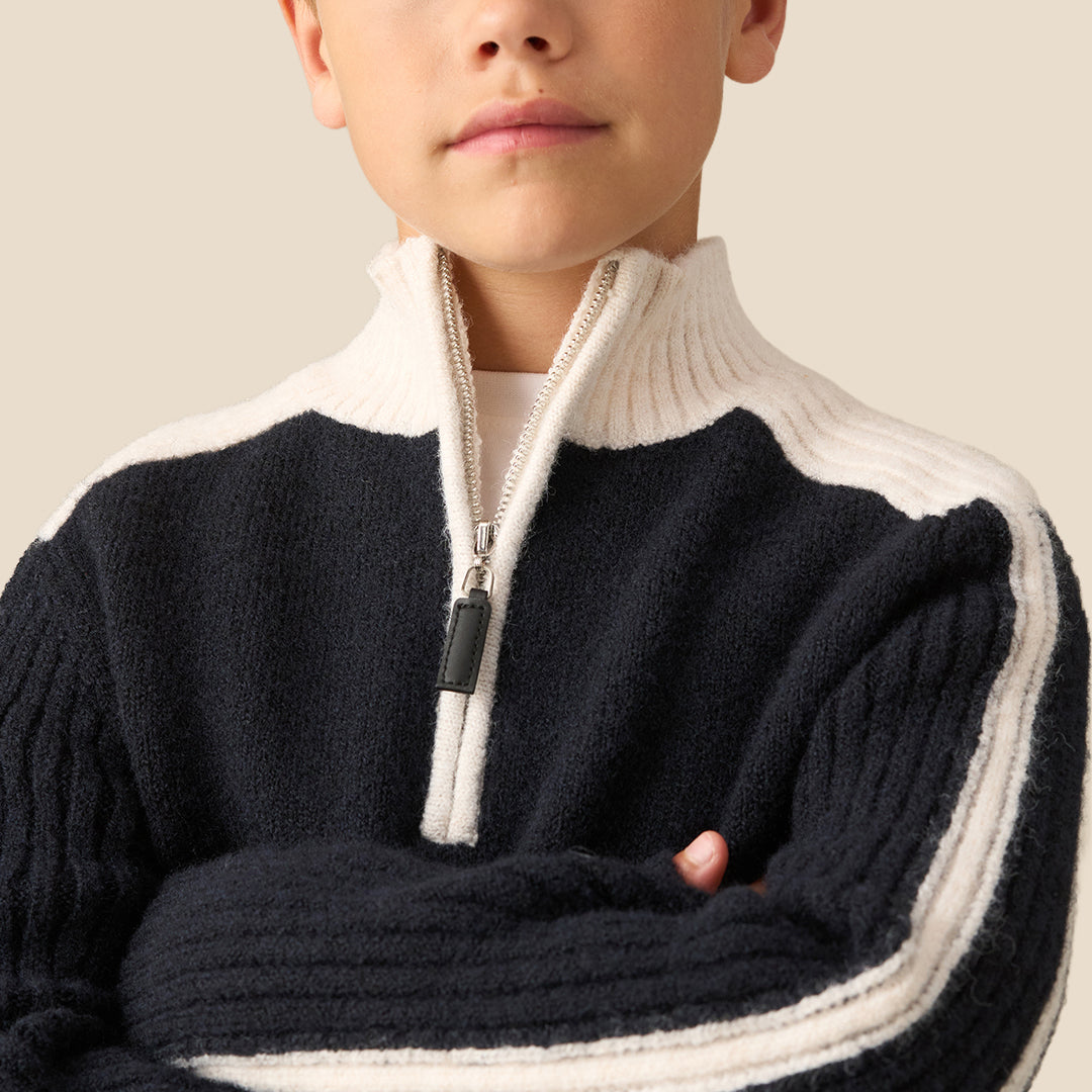 A young boy wearing a navy blue and cream knit quarter-zip sweater with white stripe details on the sleeves, paired with gray pants, against a solid beige background.