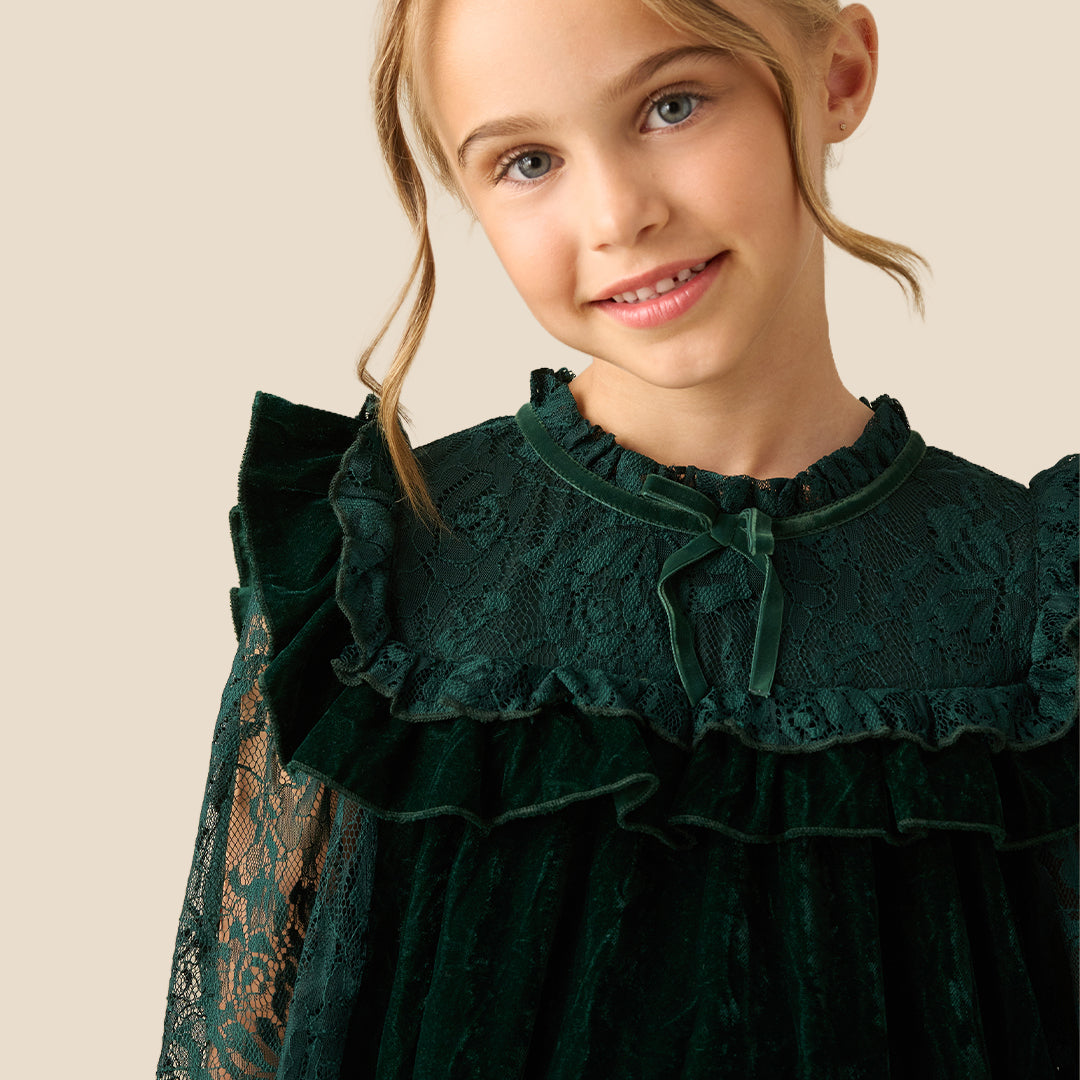 A young girl wearing a dark forest green velvet dress with lace sleeves and a ruffled high neckline, photographed against a solid beige background.