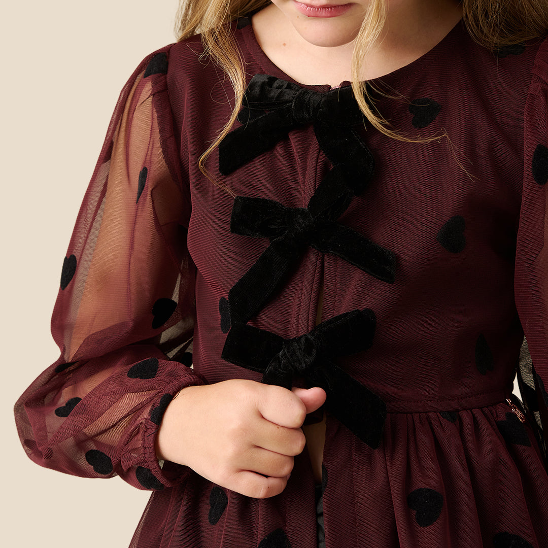 A young girl with blonde hair in a high ponytail is wearing a dark burgundy sheer long-sleeve blouse with black heart and bow details, paired with a black skirt, against a solid beige background.