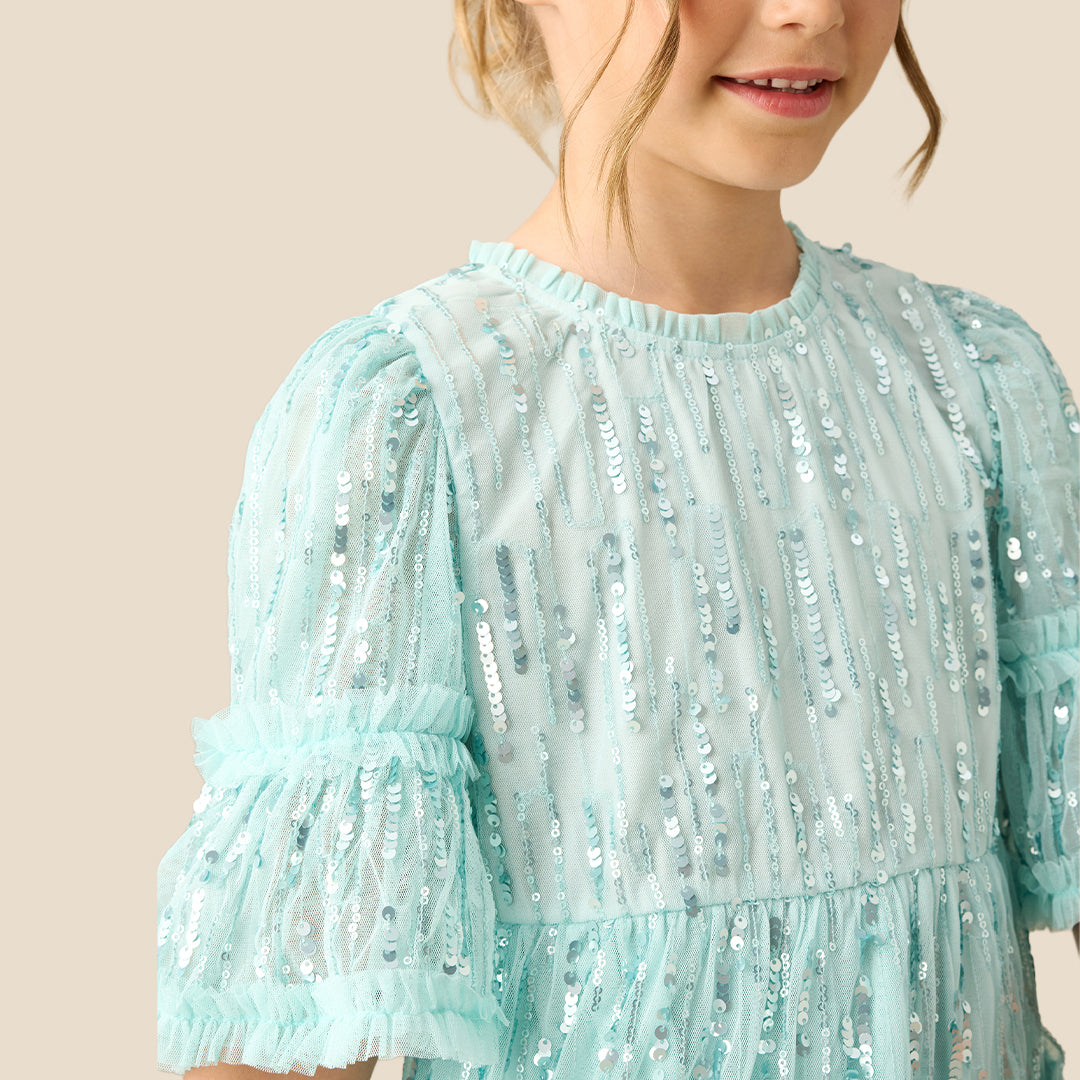A young girl with a long brown ponytail wearing a light mint green sequined tiered dress, standing against a solid beige background.
