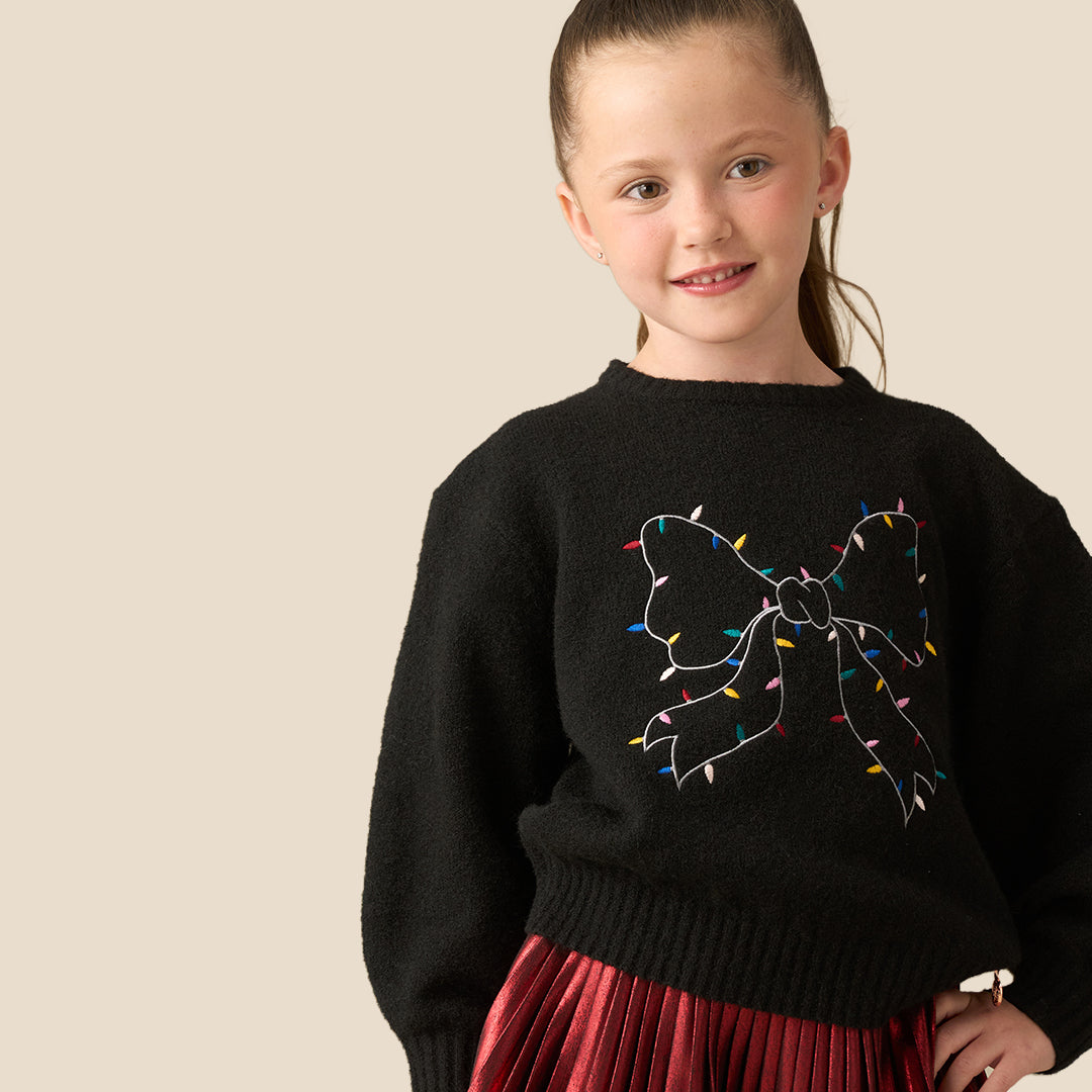 A young girl with her hair in a ponytail is wearing a black knit sweater featuring a large bow design outlined with colorful Christmas string lights, paired with a red pleated skirt, set against a solid beige background.