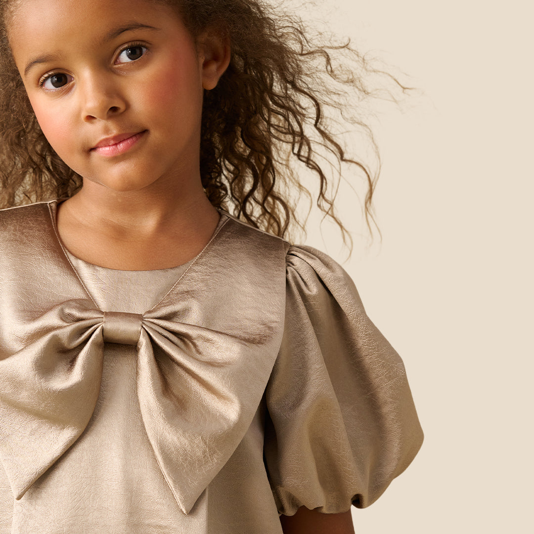 A young girl with curly brown hair wearing a champagne beige dress with puffed short sleeves and a large bow at the neckline, standing against a solid light beige background.