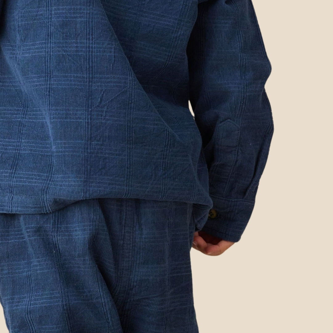 A close-up of a person wearing a dark blue textured plaid jacket and matching pants, with a hand tucked into the pants pocket, set against a solid light beige background.