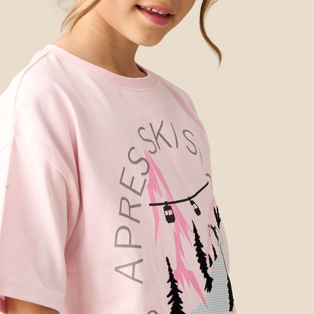 A young girl modeling a light pink graphic t-shirt with a black and pink ski design and text, paired with a gold sequined skirt, against a solid beige background.