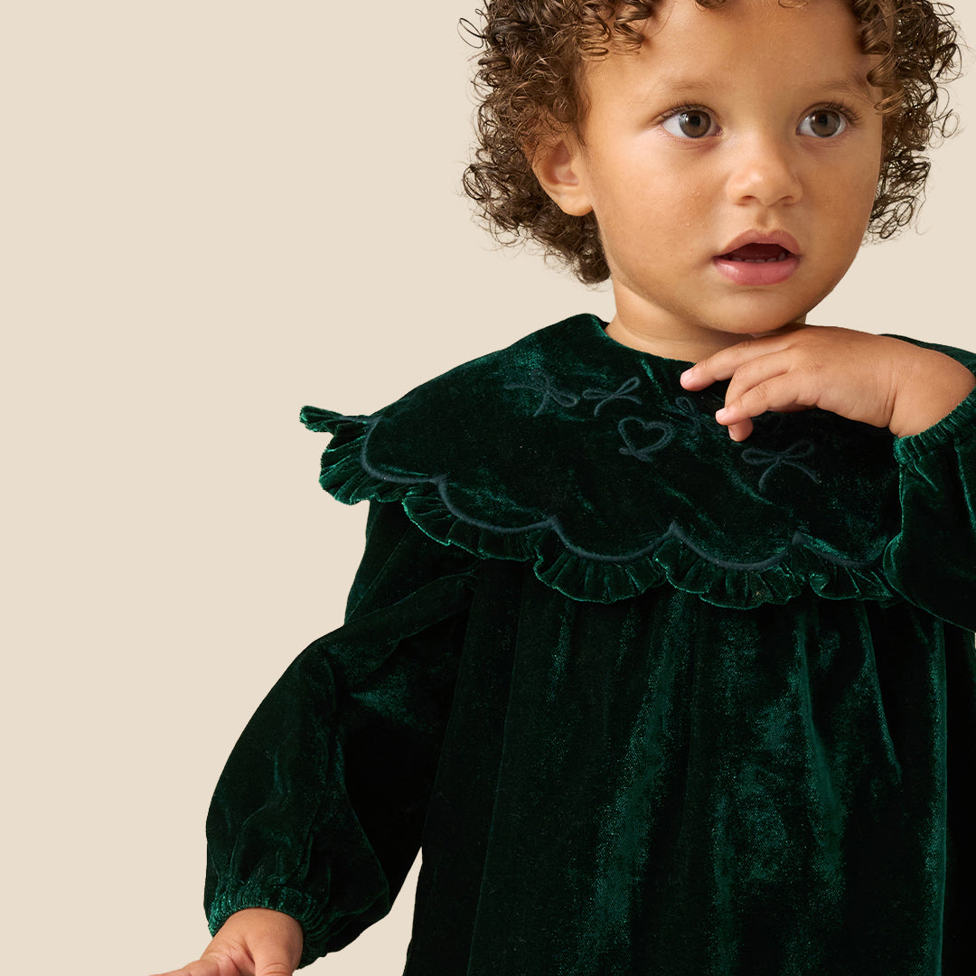 Two young children standing barefoot against a plain beige studio background, wearing matching dark green velvet dresses with wide ruffled shoulders and long sleeves.