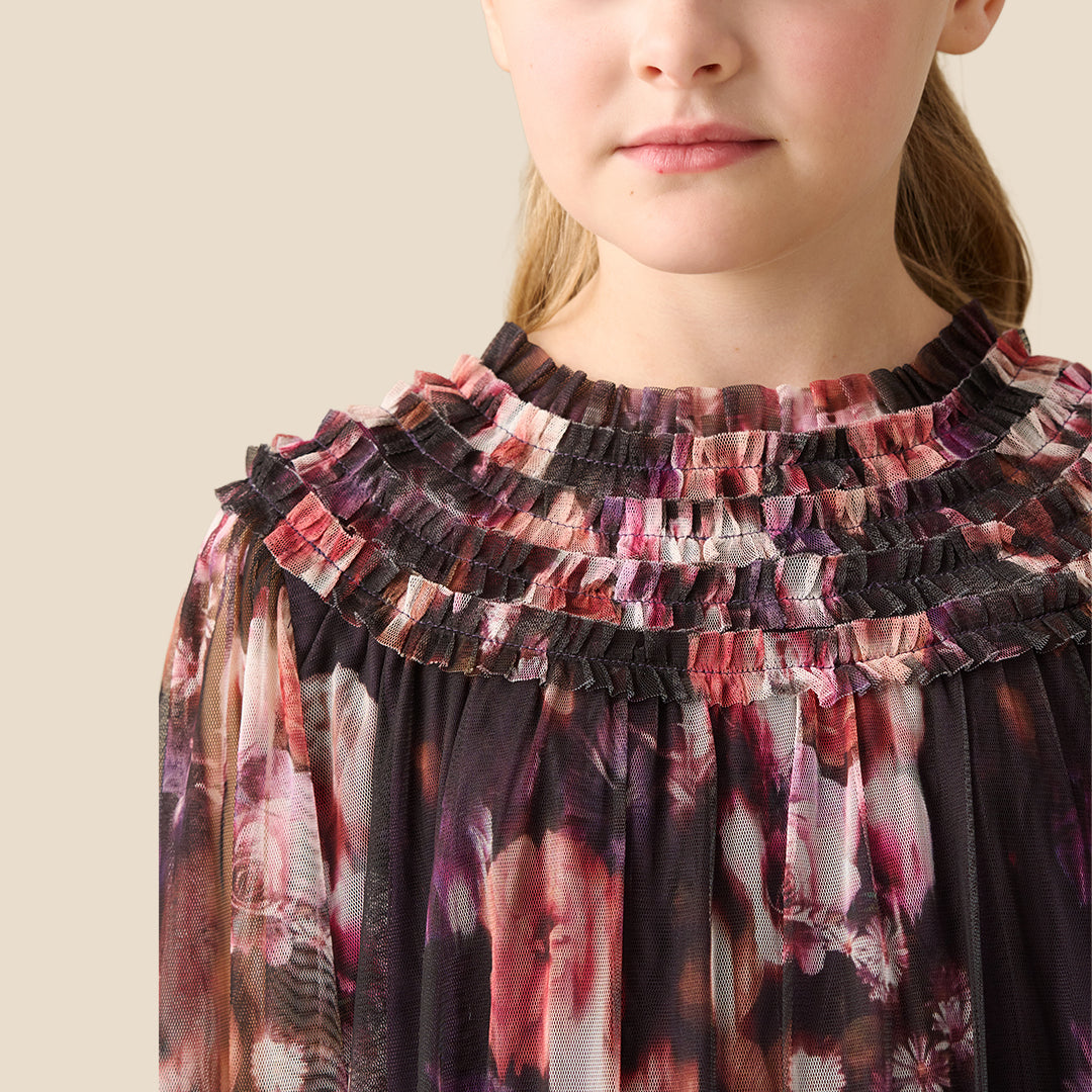 A young girl with her hair in a ponytail is wearing a long-sleeved, tiered tulle dress with a pink, red, and white floral pattern on a dark background, set against a solid beige studio backdrop.