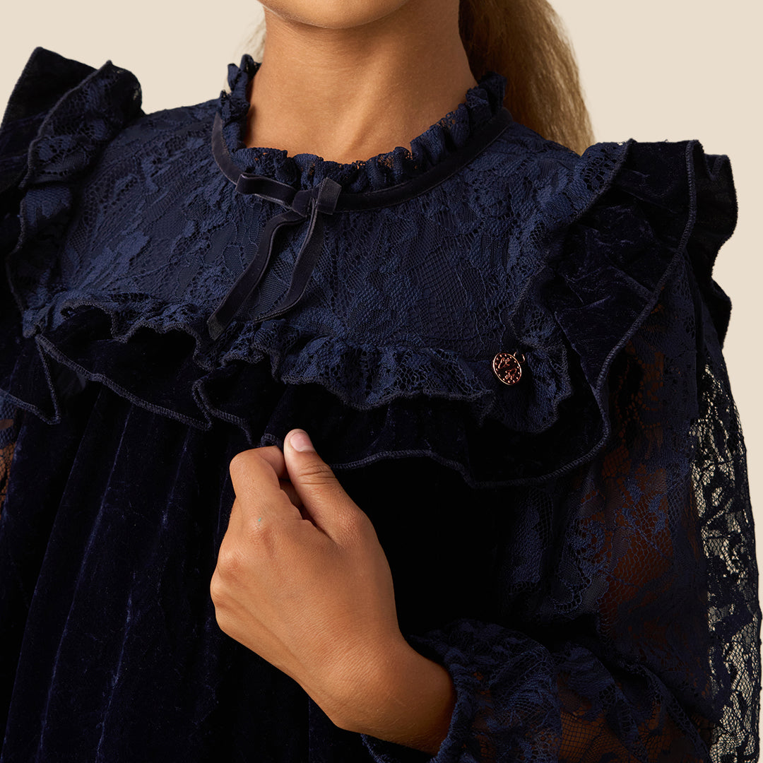 A young girl with her hair in a ponytail models a dark navy blue dress. The dress features a velvet-like texture, a ruffled collar, sheer lace long sleeves, and a tiered skirt, set against a solid beige background.